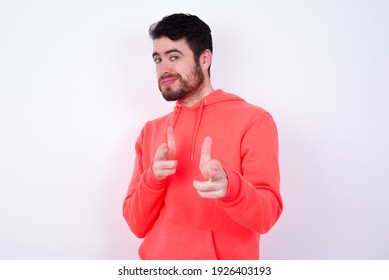 Hey you, bang. Joyful and charismatic good-looking young Caucasian bearded man wearing pink hoodie against white background winking and pointing with finger pistols at camera happily and cheeky.
