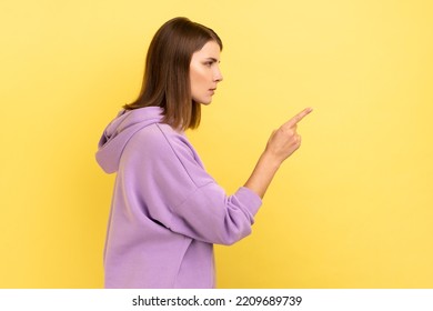Hey you, be careful. side view portrait of young dark haired woman seriously pointing finger and looking ahead warning, wearing purple hoodie. Indoor studio shot isolated on yellow background.