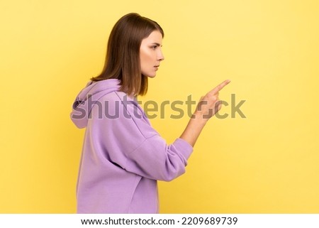 Hey you, be careful. side view portrait of young dark haired woman seriously pointing finger and looking ahead warning, wearing purple hoodie. Indoor studio shot isolated on yellow background.