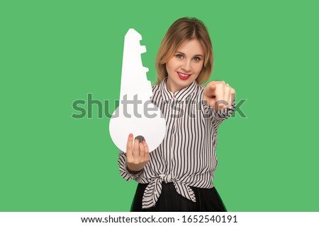 Hey you, buy house! Positive woman realtor in striped blouse holding huge key and pointing to camera, recommending real estate purchase, choosing you. indoor studio shot isolated on green background