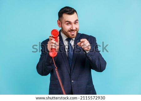 Hey you, call me Bearded young adult male wearing dark official style suit, holding retro phone handset and pointing finger to camera. Indoor studio shot isolated on blue background.