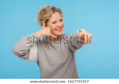 Hey you, call me! Cheerful woman with curly hair in sweatshirt doing talking on telephone gesture and pointing at camera, connection communication concept. studio shot isolated on blue background