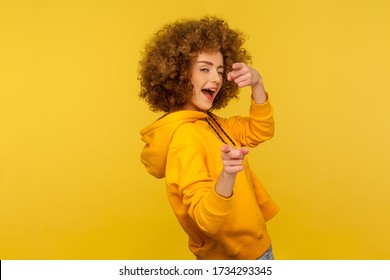 Hey you, handsome! Portrait of joyful curly-haired woman in urban style hoodie winking playfully and pointing to camera, choosing guy and flirting. indoor studio shot isolated on yellow background