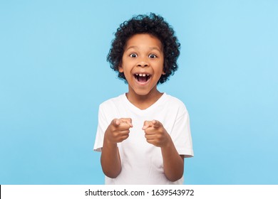 Hey You! Portrait Of Happy Little Boy With Curly Hair Pointing Finger To Camera And Laughing Loudly With Surprised Face, Teasing Making Fun Of You. Indoor Studio Shot Isolated On Blue Background