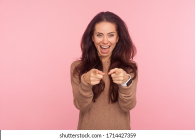 Hey You! Portrait Of Positive Happy Young Woman With Long Brunette Hair In Pullover Pointing Fingers To Camera And Laughing Loudly, Making Fun Towards You. Studio Shot Isolated On Pink Background