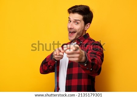 Hey, It's You. Portrait of young man in red checkered shirt pointing two index fingers at camera, posing isolated over orange background wall. Cheerful smiling guy picking, choosing and indicating