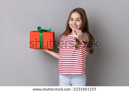 Hey, you are winner. Portrait of delighted little girl wearing striped T-shirt holding present box and pointing finger to camera with excited expression. Indoor studio shot isolated on gray background