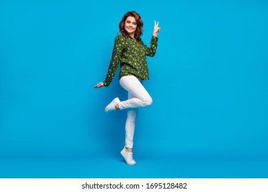 Hi guys! Full length profile photo of pretty cheerful lady good mood flirty person show v-sign symbol wear green dotted shirt white pants footwear isolated blue color background - Shutterstock ID 1695128482