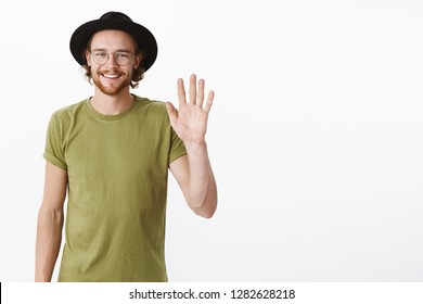 Hi nice to meet you. Portrait of handsome and charismatic young creative male newbie in office waving hand in hello gesture smiling joyfully as greeting new person in club over gray wall