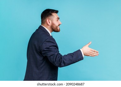 Hi, Nice To Meet You! Side View Of Friendly Smiling Handsome Man Wearing Official Style Suit Outstretching Hand, Offering Handshake On Job Interview. Indoor Studio Shot Isolated On Blue Background.