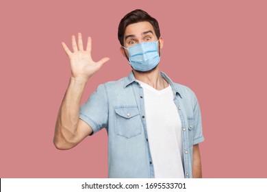 Hi, Nice To See You. Portrait Of Surprised Young Man With Surgical Medical Mask In Blue Shirt Standing And Looking At Camera With Smile And Greeting. Indoor Studio Shot, Isolated On Pink Background