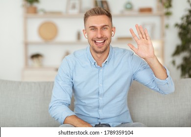 Hi. Positive Millennial Guy Waving Hello With Hand Smiling To Camera Sitting On Couch At Home, Wearing Earbuds