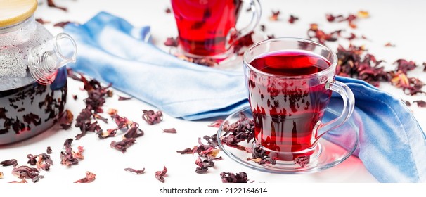 Hibiscus Tea In Glass Cup On A Blue Napkin. Cup Of Red Hibiscus Tea And Dry Hibiscus Petals On A White Background. Natural Dietary Herbal Tea