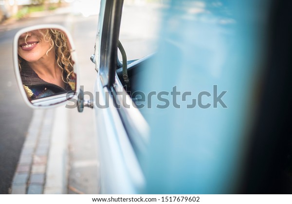 Hidden face of beautiful\
caucasian young blonde woman in the mirror of a old blue van -\
concept of travel and drive for female people - cheerful beauty\
smile and teeth