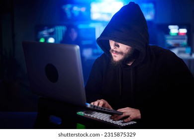 Hidden hacker in sweatshirt and hoodie hacking security systems. Unrecognizable bearded man sitting in dark room with blue neon lights. Concept of cyber attack, malware and cyber security. - Powered by Shutterstock