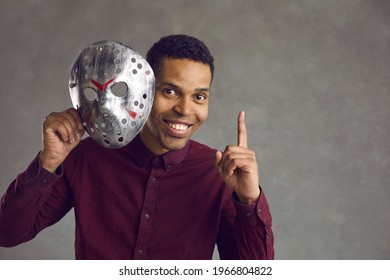 Hidden Personality: Happy Kind Handsome Young Black Man Isolated On Grey Studio Background Takes Creepy Ugly Maniac Mask Off His Face, Looks At Camera, Points Up And Smiles To Show He Can Be Different