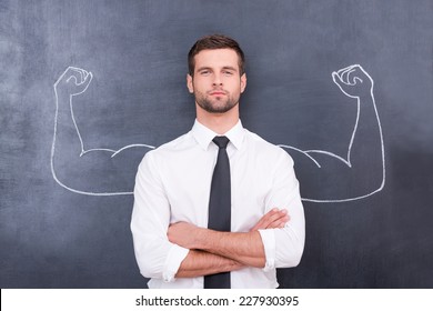 Hidden possibilities. Handsome young man in shirt and tie looking at camera and keeping arms crossed while standing against chalk drawing of muscular arms