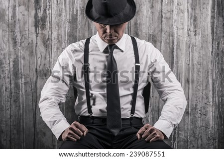 Hidden threat. Bossy senior man in gangster clothing sitting at the chair and against a wall 