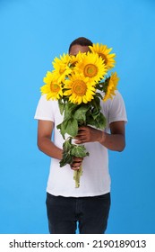 Hide Behind Bouquet Of Sunflowers Young African American Man Wearing White T-shirt And Isolated On Blue Background. Romantic Young Man With Flowers. Man With Summer Flowers. No Face Visible.