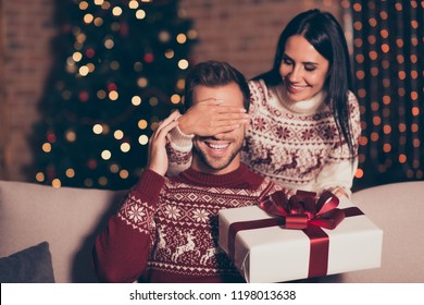 Hide and seek concept. Good-looking, attractive brunette lady in ornament sweater hold big package with bow, close eyes her man, who sit in cozy living room couch with lights garland decorations - Powered by Shutterstock