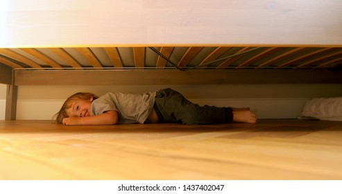 Hiding Baby Lies Laughing And Crawling Out From Under The Bed. Child Looking For Something Under The Bed.