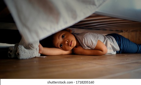 Hiding Baby Lies Laughing And Crawling Out From Under The Bed. Child Looking For Something Under The Bed.