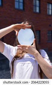 Hiding her face behind small mirror european white girl with long hair with building behind