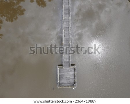 From high above, the top aerial view captures a wooden long pier extending over a serene lake on a sunny day, with the water's surface reflecting the brilliant sun, creating picturesque scenery.