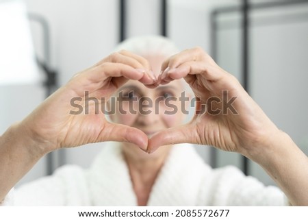 High above view of attractive old lady in bathrobe making heart symbol with her hands. Self-love caption. Beauty concept. Happiness and love