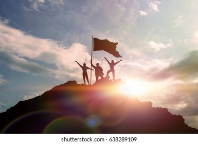 High achievement, silhouettes of three people holding a flag on the top of the mountain, hands up. A man on top of a mountain. Conceptual design. Against the dramatic sky with clouds at sunset.