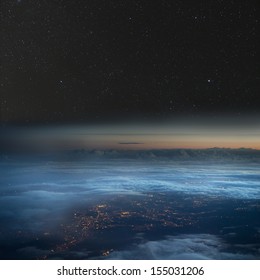 High altitude view of the Earth at night. City lights below the clouds, stars above.