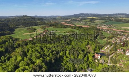 High altitude view shows patchwork farmland, dense forests, rolling hills, scattered villages, red roof houses, stone ruins, green meadows and open countryside beneath vivid blue sky in clear daylight