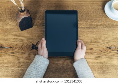 High angle above top view and cropped photo of person with modern tablet with copy space on display in hands sitting behind wooden table inside bright light interior