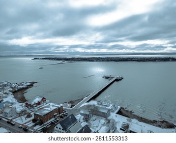 High angle aerial view captured in winter shows a snow-covered US coastal town harbor. A long pier extends into the cold, gray ocean under a dark sky.