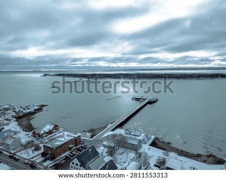 High angle aerial view captured in winter shows a snow-covered US coastal town harbor. A long pier extends into the cold, gray ocean under a dark sky.