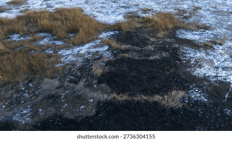 High angle aerial view of a large black charred area in a marshy field with brown reeds and snow cover during winter
