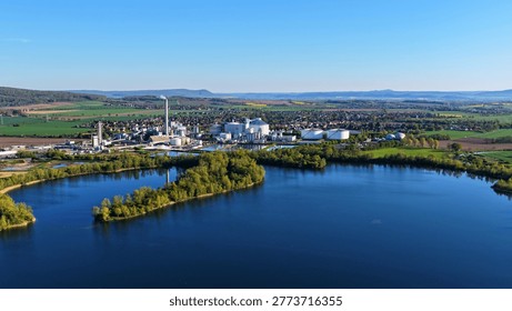 High angle aerial view of a large industrial production facility with smokestack, storage tanks, processing buildings and utility infrastructure located next to a river in a green rural landscape. Modern European manufacturing plant surrounded by farmland and residential areas, representing industry, economy, logistics, employment and environmental topics.