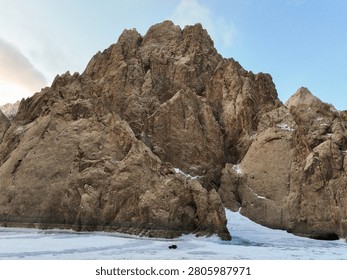 High angle aerial view of a massive, textured rock cliff face dominating the frame, with a single vehicle (appearing as a dark dot) on the frozen ice below