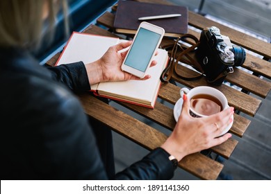High angle back view of faceless lady sitting in cafeteria with tea and notebook with pen and camera with book at table while checking cellphone