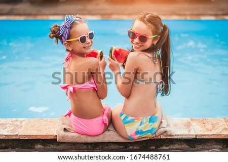 High angle back view of happy friends in sunglasses and colorful swimsuits with slices of fresh tasty watermelon looking, over shoulder at camera and smiling while sitting on poolside pool