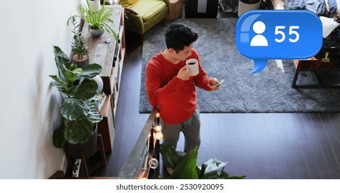 High angle of a Caucasian man drinking coffee while texting on his phone in the living room. Above him in the foreground is a digital image of a message bubble with a follower icon increasing in - Powered by Shutterstock