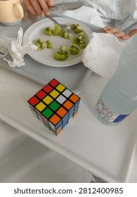 High angle, close up shot focused on a colorful 4x4 Rubik's cube on a hospital bed food tray. In the background, partially in focus, are a child's hands eating kiwi from a plate, a cup, and a water bottle.