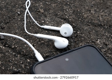 High angle close up view of a pair of classic white wired earphones plugged into a smartphone, resting on a rough concrete or asphalt outdoor surface