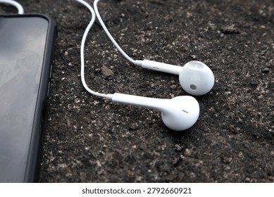 High angle close up view of a pair of white wired earphones plugged into a smartphone, resting on a dark, rough concrete or asphalt outdoor surface