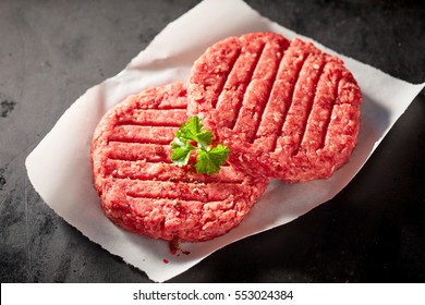 High Angle Close Up View Of Two Raw Beef Hamburger Patties Topped With Sprig Of Fresh Parsley And Resting On Square Of Wax Paper On Dark Gray Stone Counter Surface