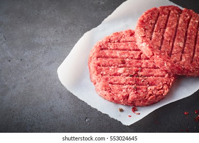 High Angle Close Up View Of Two Raw Beef Hamburger Patties On Square Of Wax Paper Resting On Dark Gray Stone Textured Counter Surface With Copy Space