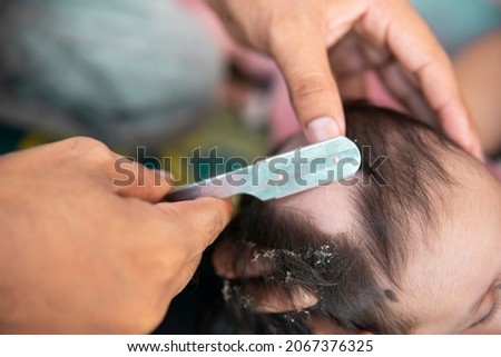 High angle close-up of Indian, newborn baby getting a first haircut with a razor blade.  