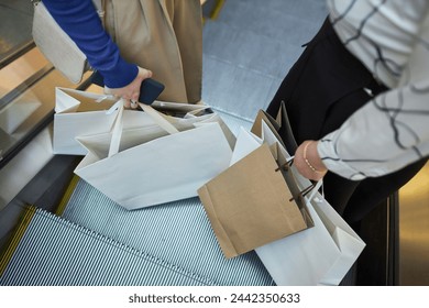 High angle closeup of two unrecognizable women holding shopping bags while standing on escalator in mall