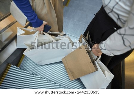 High angle closeup of two unrecognizable women holding shopping bags while standing on escalator in mall