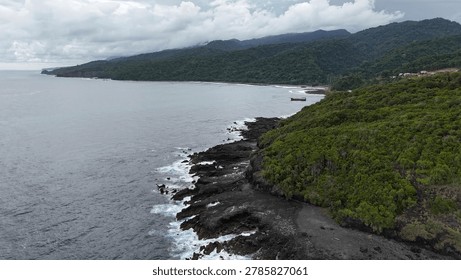 High angle coastal view of a continuous green mountain range meeting the dark volcanic rocky shore under overcast sky, moody nature background with copy space.